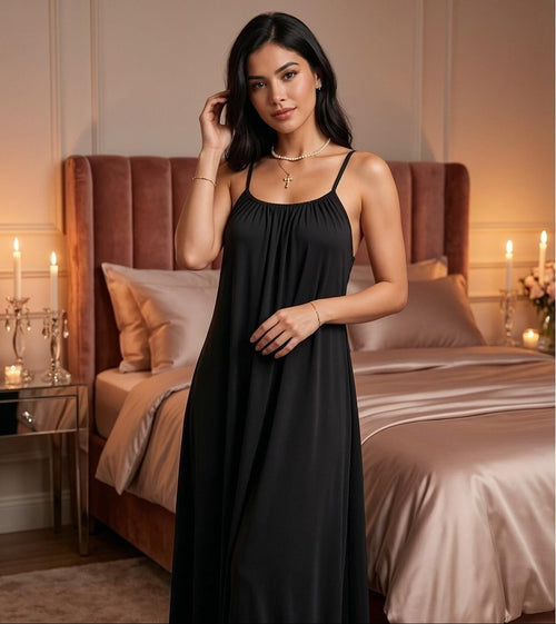Model wearing a black backless maxi dress and silky lounge gown by Soft Secrets in a luxury bedroom setting.
